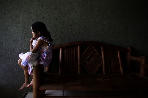 Ein Mädchen sitzt mit einem Kuscheltier im Arm auf der Lehne einer Bank in einem dunklen Raum