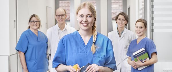 Eine blonde, junge Pflegekraft steht im Gang der Klinik und blickt in die Kamera. Hinter ihr stehen ein Arzt, eine Ärztin und zwei weibliche Pflegekräfte und blicken ebenfalls in die Kamera.