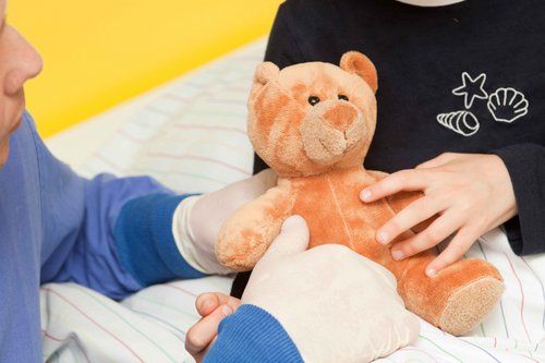 Ein Patient hält einen Teddybären, eine Pflegerin sitzt daneben