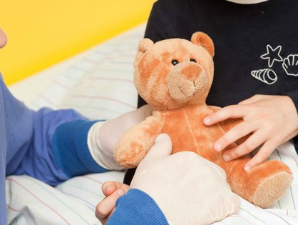 Eine medizinische Fachkraft sitzt am Bett eines Kindes, das einen Teddybär im Arm hält