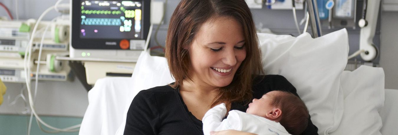 Eine Mutter sitzt im Krankenbett und hält lächelnd ihr Baby im Arm an 
