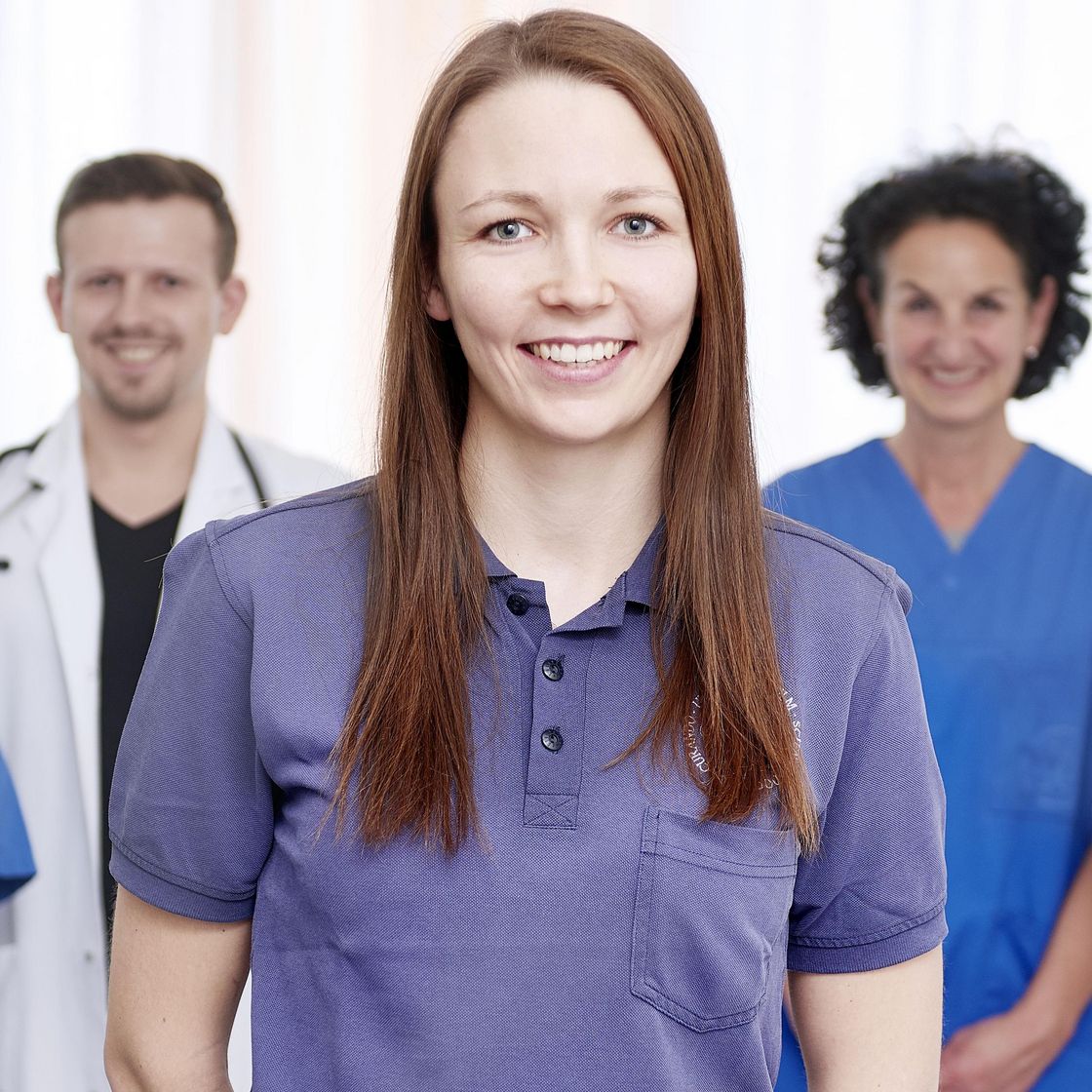 Im Vordergrund eine junge Physiotherapeutin im blauen Shirt, im Hintergrund Ärzte und Pflegekräfte