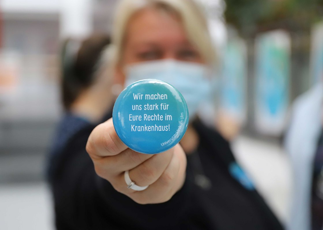 Eine Mitarbeiterin hält einen Button mit der Aufschrift "Wir machen uns stark für eure Rechte im Krankenhaus" in die Kamera.