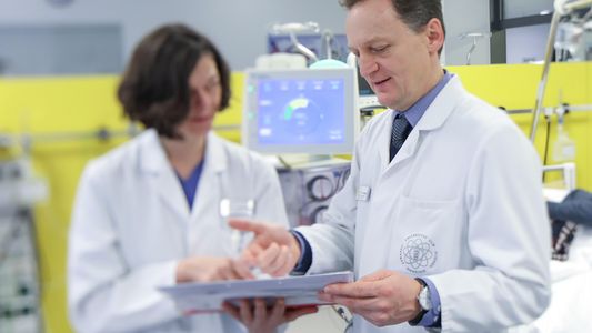 Ein Arzt und eine Ärztin blicken auf ein Blatt Papier und unterhalten sich. Im Hintergrund ist ein Monitor zu sehen. 