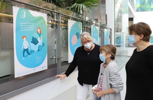 Eine Patientin betrachtet mit einer Therapeutin und ihrer Mutter die neue Ausstellung in der Kinderklinik.