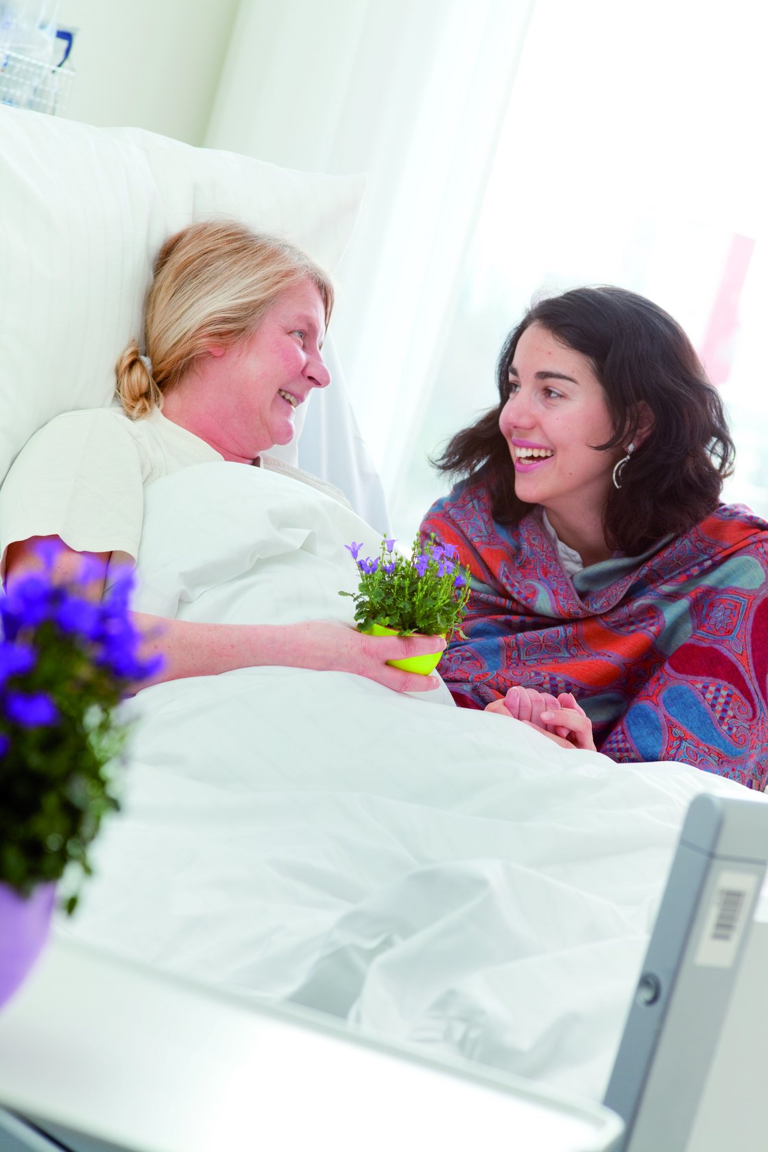 Eine Patientin im Krankenbett lacht eine Besucherin an 